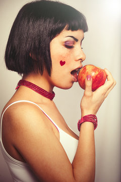 Eating a red apple. Young woman love for fruits. A young girl about to eat a red apple. The girl has a heart sticker on the cheek. Freckles and blacks bobbed hair. On clear graduated background.