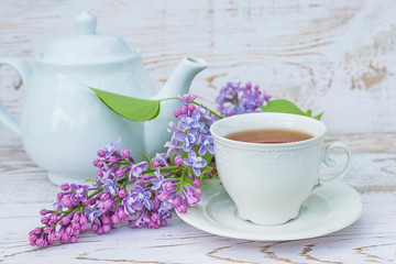 Spring tea time still life in retro style. White cup of tea, tea pot and bunch of purple lilac on white pant wooden background