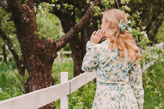 Back view of young attractive woman in white retro dress posing on green countryside garden. Beautiful stylish blonde hippie girl with bow in hair standing in park. Girl enjoy summer nature outdoors