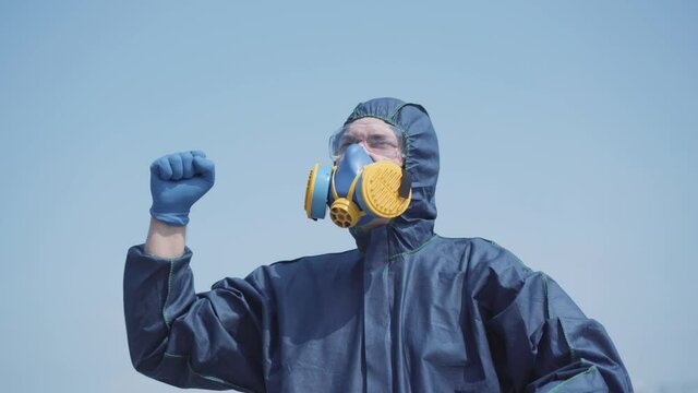 Stressed Caucasian man in respirator and biohazard suit shouting and showing strength gesture at sky. Young guy tired of global pandemic or chemical pollution yelling in nowhere outdoors on sunny day.