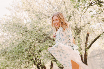 Fototapeta na wymiar Portrait cheerful young hippie girl with wavy hair have fun and smile while walk in green sunny park. Beautiful happy blonde woman in stylish floral dress dancing on countryside garden. Enjoy nature