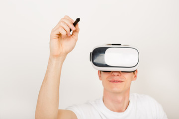 Male using virtual reality headset interacts with augmented things orienting in three dimensional space while sitting on his desk with keyboard and laptop on white background.