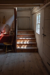 Interior of a very old fishing shack, light streaming through window onto stairs, table and chair, vertical aspect