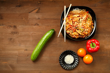 Black plate with funchose and chicken with vegetables under pad Thai sauce on a wooden table and fresh vegetables.
