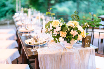 Festive outdoor wedding celebration table covered with natural tablecloth and beautifully neatly served with sets of dishes, glasses, appliances and flower arrangements on a green background.