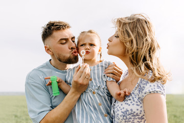 Happy young family close-up portrait outdoors. Mother, father and little daughter making with soap bubbles.