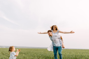 Young couple of man and woman playing in an open field, outdoors, on clean white background. Man gives a piggyback riding to woman, laughing. Woman with arms wide open, like flying.