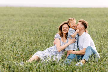 Fototapeta premium Happy young family spending time outside, hugging each other. Woman, man and a little girl on a summer day, sitting in a wheat green field.