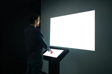 Man using electronic kiosk and looking at white blank large interactive wall display in dark room...