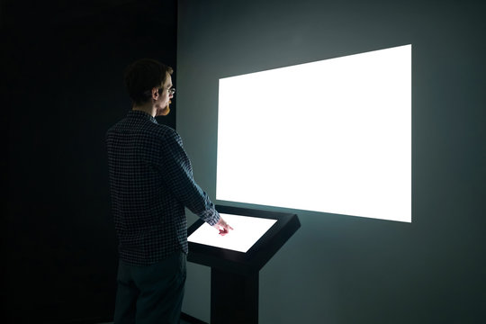 Man Using Electronic Kiosk And Looking At White Blank Large Interactive Wall Display In Dark Room Of Modern Technology Exhibition. Mock Up, Futuristic, Template, Education, Technology Concept