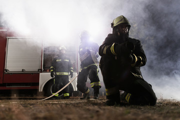 Team of professional firefighters holding water hose in front of a firetruck with smoke in the air. Half silhouette.
