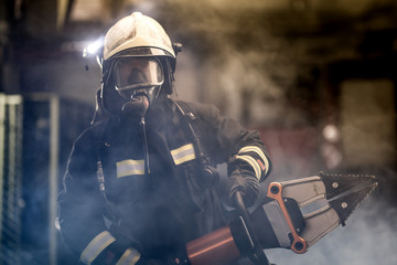 Obraz premium firefighter portrait wearing full equipment, oxygen mask, and power hydraulic cutting tool, smoke and fire trucks in the background.