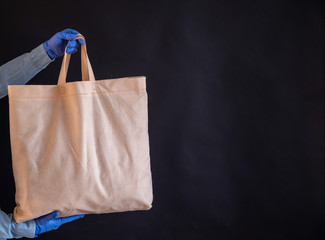 A bag of cotton fabric on a black background. The courier holds a canvas cloth bag without plastic packaging on a black background. Safe delivery to the epidemic. A closeup of hands in gloves.