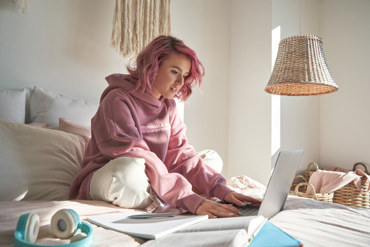 Hipster teen girl school student with pink hair wearing hoodie using laptop computer sitting in bed distance elearning online learning course for exam search remote online classes in bedroom at home.