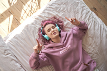 Happy funny teen girl with pink hair wear headphones lying in comfortable bed listening new pop...