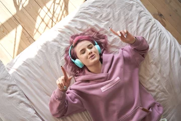 Poster Happy funny teen girl with pink hair wear headphones lying in comfortable bed listening new pop music enjoying singing song with eyes closed relaxing in cozy bedroom at home. Top view from above. © insta_photos