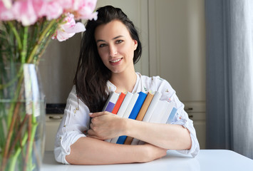 Indoor horizontal portrait of smiling business woman posing at home with her reading stack of books. Smart female student holding a stack of books. Lifestyle, business, and education concept