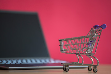 Online shopping concept. Mini grocery cart and laptop on a pink background. Small cart and personal...