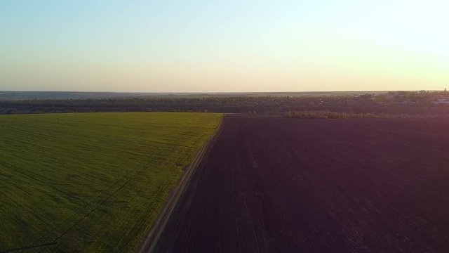 Sunset over agricultural famland. Aerial photography: countryside, green meadow and plowed black field, camera flies along border of the field and the earth, at sunset in spring, summer.