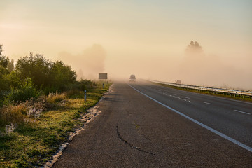 Fog on highway. Trees in haze. green bushes and wildflowers along road. Asphalt road with separate lane and bumper. Car bus on road with headlights on.