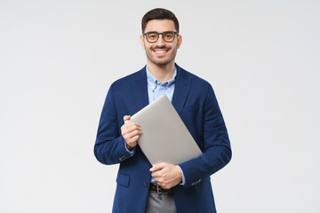 Smiling business man looking at camera through trendy glasses, holding closed laptop next to chest...