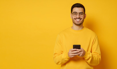 Horizontal banner of young man in eyeglasses, smiling happily while looking at camera after communication with friends on phone, isolated on yellow background, copy space on left