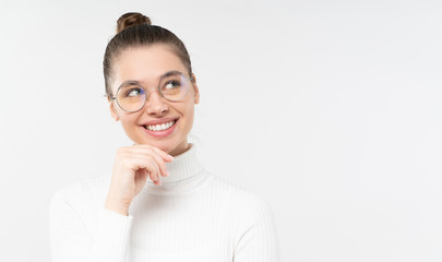 Horizontal banner of young female wearing eyeglasses and white sweater, holding chin, dreaming of future with smile, isolated on gray background with copy space