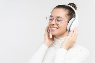 Naklejka premium Pretty girl in wireless headphones, pleased with quality sound of music of her favorite songs, smiling with eyes closed, isolated on gray background, copy space
