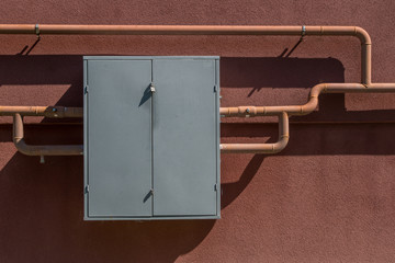 Grey metal closed on padlock control cabinet fixed to brown facade of residential apartment building. Through cabinet pass metal pipes of the gas pipeline, attached to the wall with round clamps.