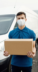 Portrait of young courier in mask standing near delivery car and holding carton box