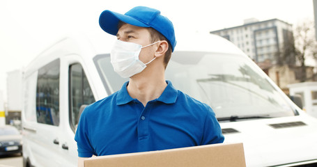 Naklejka na ściany i meble Portrait of young courier in mask standing near delivery car and holding carton box