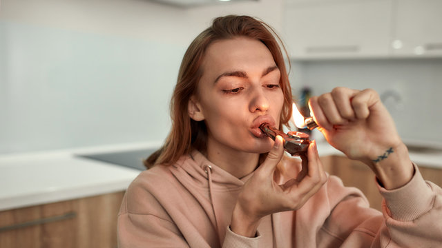 Stress Away. Young caucasian woman lighting cannabis in the bowl of metal pipe while sitting in the kitchen. Buds in a plastic bag and red marijuana grinder on the table