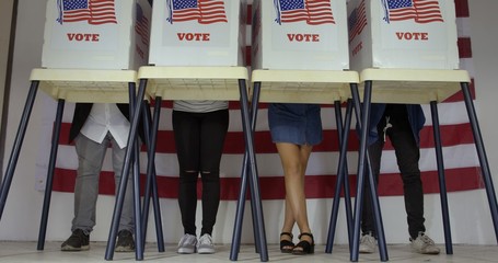 MS Legs of two men and two women of various demographics as they stand at voting booths and complete ballots in polling station with US flag.