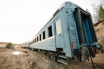 Fototapeta na wymiar A blue, destroyed train stands on rails with broken windows in the middle of a field around a sandy area