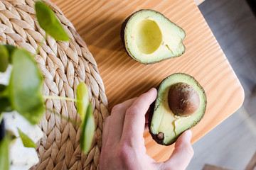 Mano cogiendo un aguacate cortado por la mitad sobre una mesa de madera y una planta verde