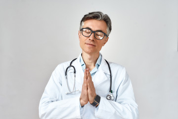 Senior male doctor wearing white coat and glasses, with stethoscope around neck keeping eyes closed and praying for help while standing against grey background