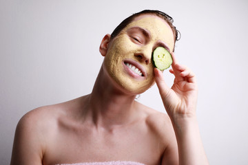 Young caucasian woman with a face mask on holding a cucumber slice in front of one eye. Beauty and...