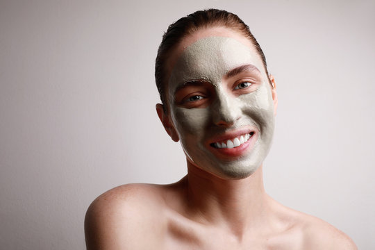 Close-up portrait of happy woman with clay facial mask with cute smile isolated on grey wall. Beauty and skin care.