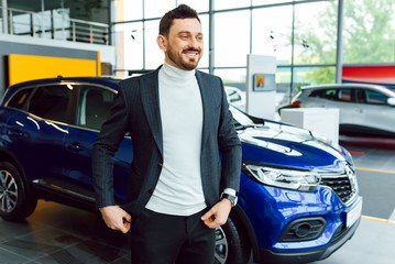 Fototapeta na wymiar Handsome bearded buyer in casual wear in dealership, guy looks on camera while standing near car with crosed arms
