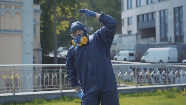 Funny workout of man in respirator and chemical suit outdoors. Portrait of young Caucasian sportsman stretching on sunny day during Covid-19 pandemic or air pollution. Fun, humor, lifestyle.