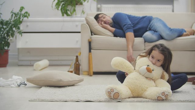 Daughter playing while drunk dad sleeping on sofa behind in 4k video.
