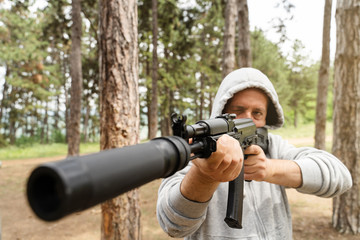 Front view of caucasian man wearing hood holding rifle semi automatic gun with silencer in the...