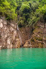 Wushan, Chongqing, China - May 7, 2010: Mini Three Gorges on Daning River. Fine lang white waterfall over brown rock cliff as shoreline behind emerald green water. Foliage on top.