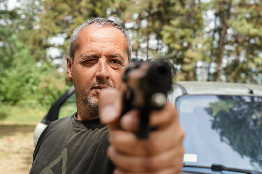 Front view head and shoulder portrait of mature caucasian man aiming hand gun holding to the camera ready to shoot in sunny summer day serious threatening murderer or shooting outdoor in nature woods