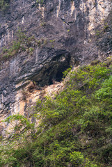 Fototapeta na wymiar Wushan, Chongqing, China - May 7, 2010: Mini Three Gorges on Daning River. Closeup of ancient ancestral cemetery black cave high up in brown-black cliff with green foliage around. 