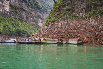 Wushan, Chongqing, China - May 7, 2010: Mini Three Gorges. ten covered tourist sampans with docked against brown cliff rock  behind emerald green water. Green foliage. Flags add color.