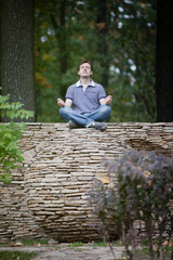 Fototapeta premium Man is meditating on stone ball in forest. Outdoor meditation surrounded by nature in yoga pose. Maintaining peace and quiet. Inner harmony. Time alone with yourself. Self-isolation. Survive stress