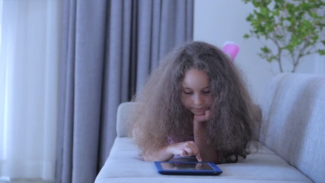 Curious cute girl preschool child using a digital tablet technology device looks telephone.Plays in the phone sitting on the sofa alone.Children tech addiction concept play game at home.