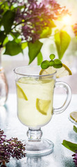 Refreshing cold yellow lemonade in a transparent cup and lilac in a vase are on the table