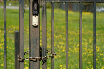 Metal gate with a bell closed on a chain and a lock, on a blurry background of a blossoming lawn. Quarantine.
